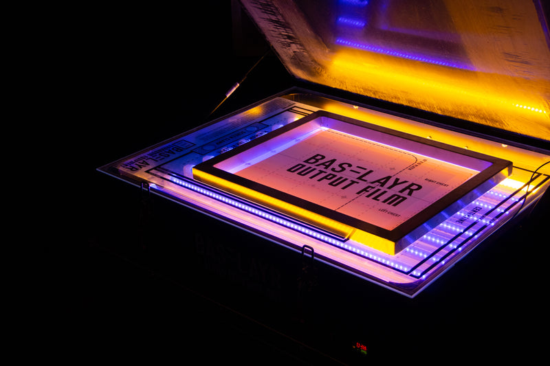 Screen exposure unit with a glowing orange and blue light, holding a screen and Baselayr output film in place. The darkroom setup emphasizes the precision and technology used to create sharp, detailed stencils for screen printing.