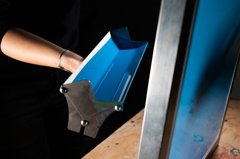 Person holding a scoop coater filled with bright blue emulsion, positioned next to a screen ready for coating. The setup highlights the preparation stage of screen printing, where precision and even coverage are key for creating quality stencils.