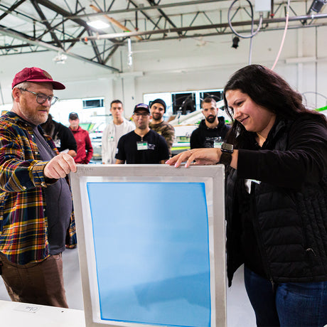 Group of screen printing enthusiasts gathered in a workshop, watching a hands-on demonstration. Two participants hold a screen frame while others observe, creating a collaborative learning atmosphere filled with creativity and technique-sharing