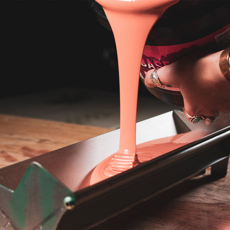 Close-up of a vibrant peach-colored ink being poured from a container onto a screen printing scoop coater. The smooth flow and rich texture of the ink highlight its quality, ready to be spread evenly for precise screen printing.