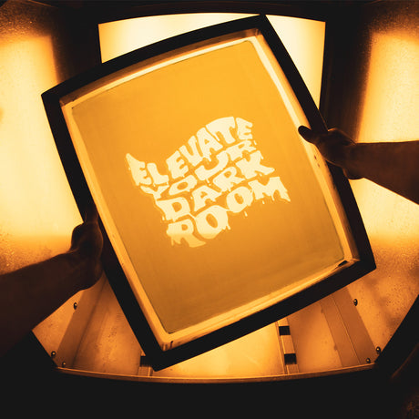 Hands holding a screen with the phrase 'Elevate Your Dark Room' illuminated against a warm, orange backdrop. The bold, dripping text design shines through, capturing the process of screen exposure in a darkroom setting, essential for creating crisp, detailed prints.