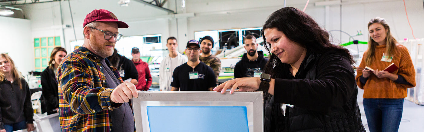 Group of screen printing enthusiasts gathered in a workshop, watching a hands-on demonstration. Two participants hold a screen frame while others observe, creating a collaborative learning atmosphere filled with creativity and technique-sharing.