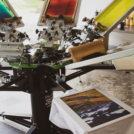 Riley Hopkins 360 screen printing press set up with multiple screens in vibrant colors, ready for multi-color printing. A printed shirt with a detailed, colorful design is positioned on the platen, showcasing the precision and versatility of the screen printing process.