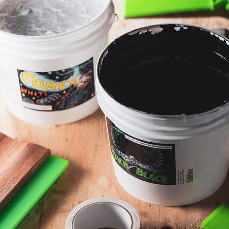 Two buckets of screen printing ink labeled 'Comet White' and 'Pitch Black' on a wooden workbench, with bright green squeegees nearby. The vivid labels and contrasting ink colors highlight essential supplies for creating sharp, high-quality prints.