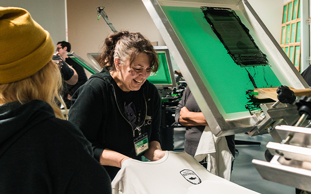 Group of people screen printing in a lively workshop setting. A smiling woman prepares a white t-shirt under a screen with black ink, capturing the hands-on, creative, and collaborative spirit of screen printing.