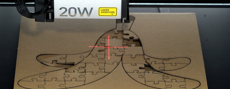 Close-up of a 20W xTool laser cutter creating a banana-shaped jigsaw puzzle on a cardboard sheet. The red laser crosshairs emphasize precision cutting, producing clean, intricate puzzle pieces with smooth edges.