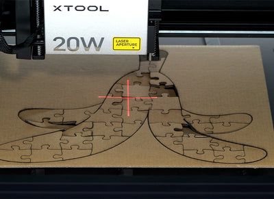 Close-up of an xTool 20W laser cutter engraving a banana-shaped jigsaw puzzle onto a piece of cardboard. The red laser crosshairs highlight the precision of the cutting process, creating clean, intricate puzzle pieces