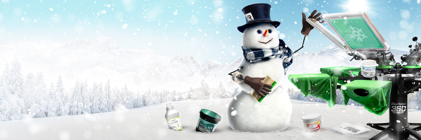 Festive winter scene featuring a cheerful snowman wearing a top hat with an 'R' logo, a scarf, and gloves, holding a squeegee. A Riley Hopkins 360 screen printing press is set up beside the snowman with a green shirt being printed with a snowflake design. Screen printing supplies, including inks and cleaning products, are displayed in the snowy foreground. The background showcases a snowy mountain landscape with falling snow and frosty pine trees, creating a holiday-themed atmosphere.