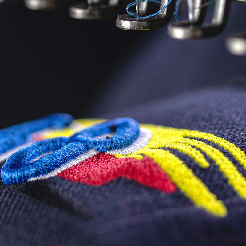 Close-up of a colorful embroidery design being stitched on fabric, with vibrant blue, red, and yellow threads. The precision and texture of the stitching highlight the quality and detail achievable with professional embroidery machines