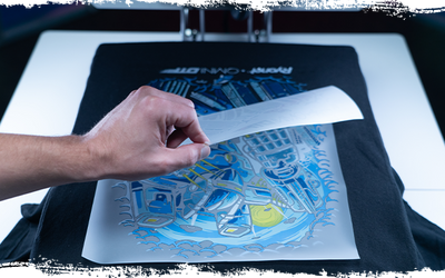 Hand lifting a transfer sheet to reveal a detailed, vibrant design on a black t-shirt, positioned under a heat press. The intricate artwork displays crisp lines and bright colors, showcasing the effectiveness of high-quality heat transfers.