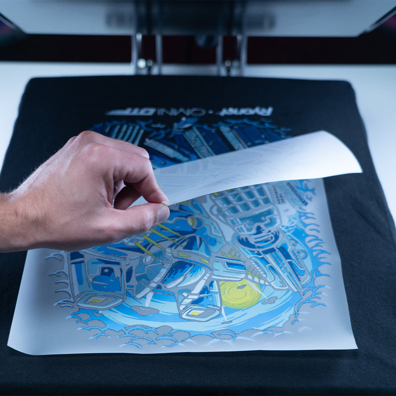 Hand carefully peeling back a transfer sheet to reveal a vibrant, detailed design on a black shirt, set up under a heat press. The intricate graphic showcases crisp lines and bold colors, demonstrating the effectiveness of high-quality heat transfers